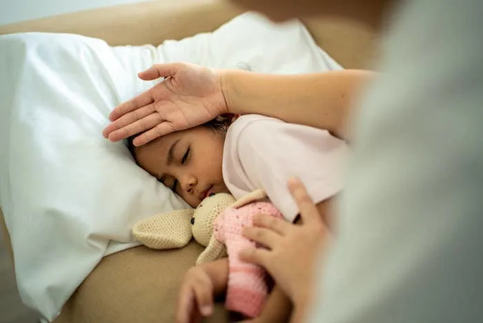 Child sleeping in bed, holding a stuffed toy. An adult hand gently checks the child's forehead