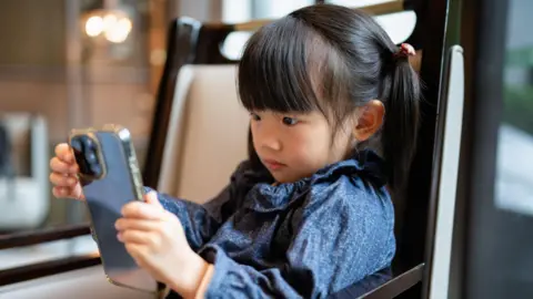 Getty Images A stock photo of little girl with pigtails looks intently at her smartphone. She is sitting , seated in a modern, warmly lit indoor environment an wearing a patterned blue top.