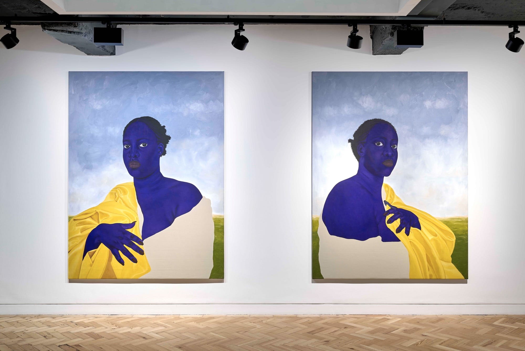 An installation view of two paintings by Stacy Gillian Abe, each featuring a blue-toned woman wrapped in fabric with a blue sky in the background