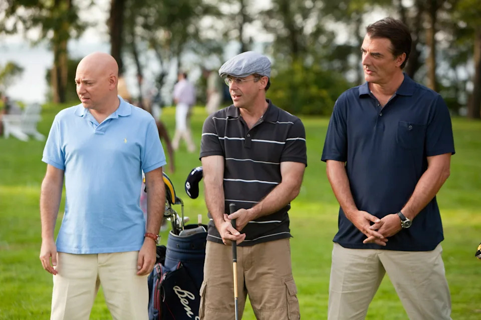 Three men stand on a golf course, wearing casual golfing attire. One holds a golf club. Trees and people are in the background