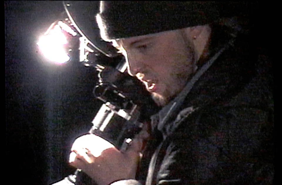 Person holding a video camera under bright light, wearing a beanie and jacket, focused on capturing footage