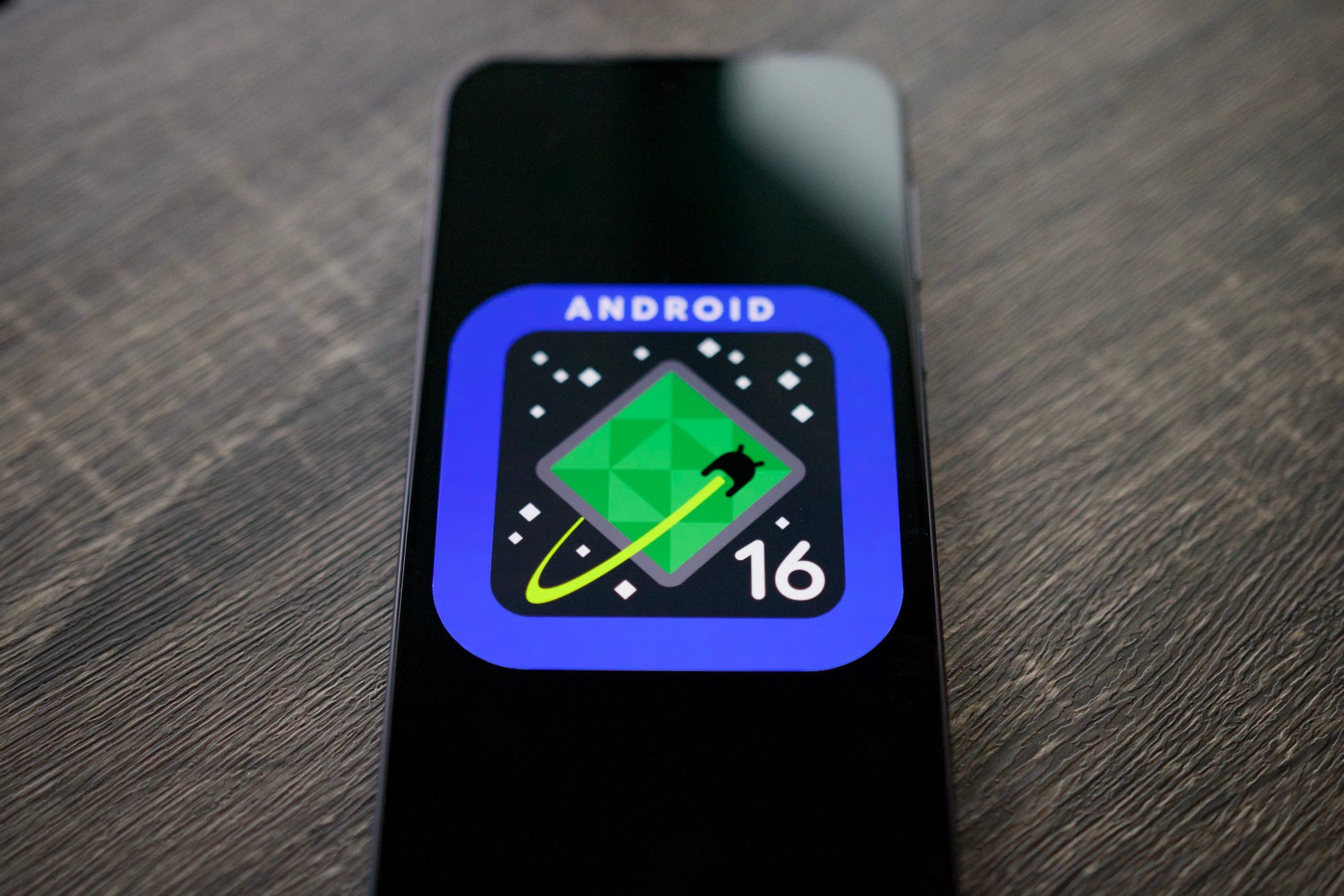 The Android 16 logo on a smartphone, resting on a shelf.