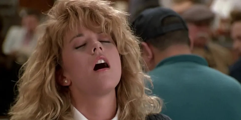 Person with curly hair appears to be mid-sentence, seated in a crowded indoor setting