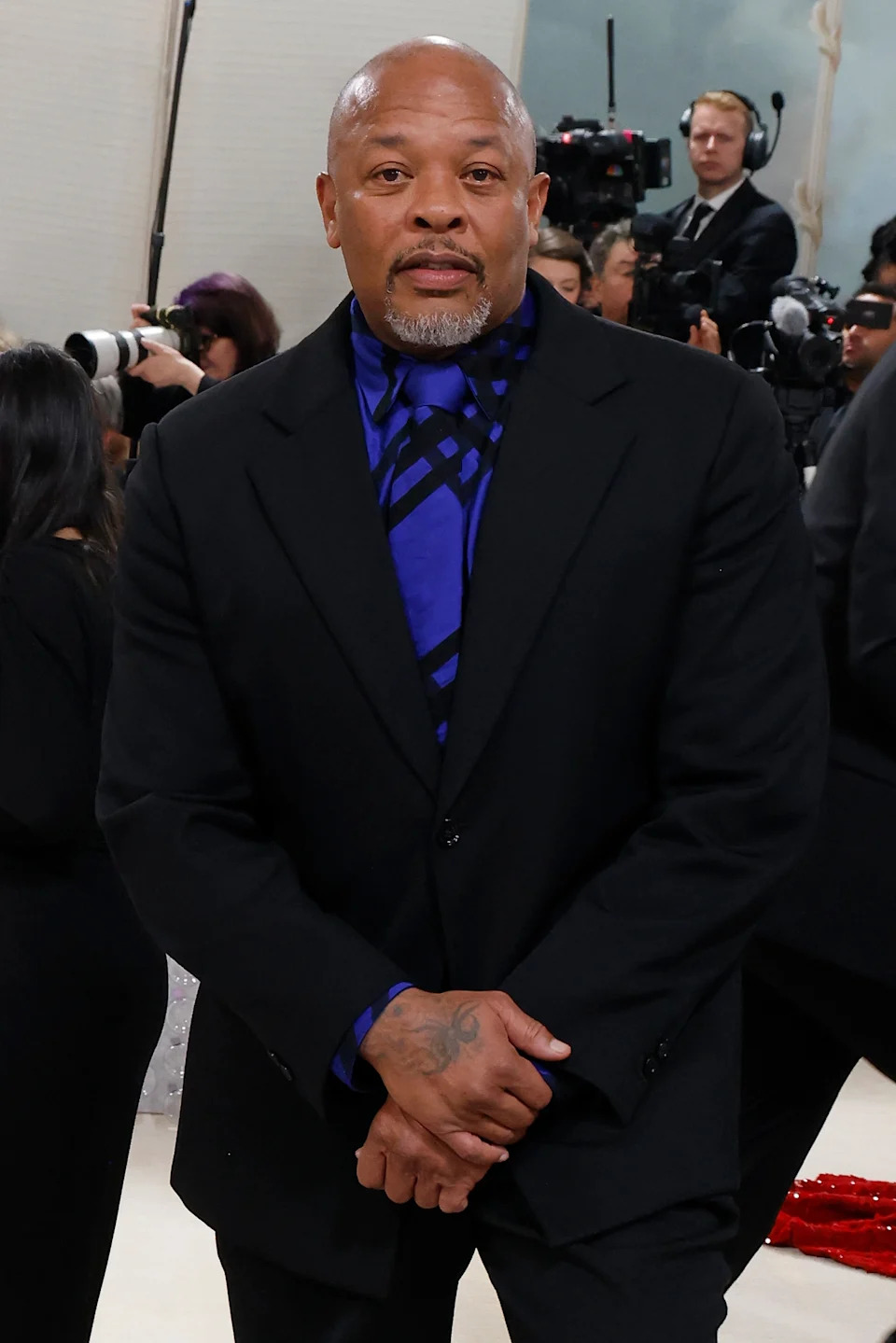 A man on the red carpet wears a sleek black suit with a patterned shirt, surrounded by photographers