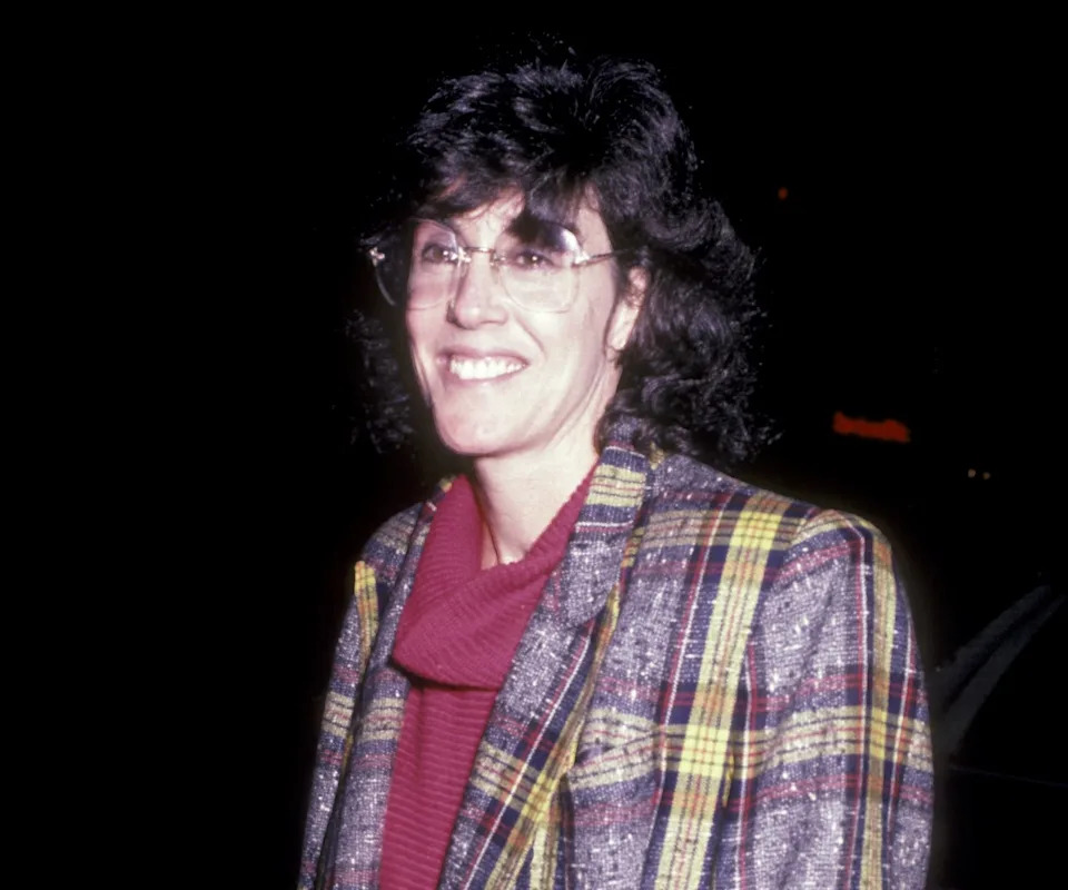 A person wearing a plaid jacket over a turtleneck and skirt, smiling, standing outdoors at night