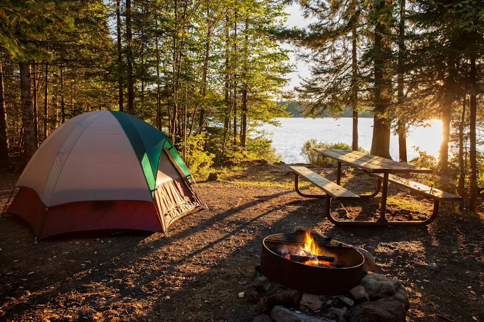 Tent and campfire near a lake, surrounded by trees. Picnic table in the background. Sunlight filters through the forest