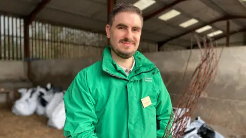 BBC A man with brown hair and facial hair smiles as he stands in a barn, holding a sapling. Large white bags containing saplings can be seen behind him. He is wearing a green, Forestry England branded coat, and a name badge.