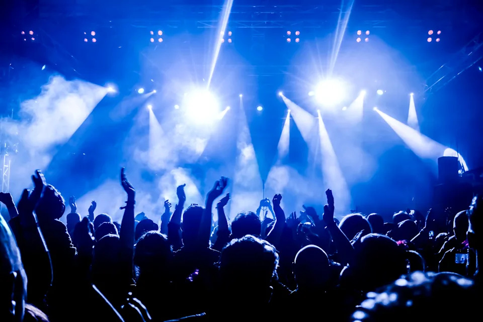 Concert crowd cheering, hands raised, silhouetted against bright stage lights during live performance