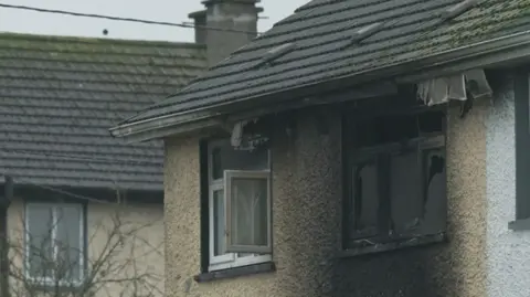 RTÉ The camera is zoomed in on the top two windows of a light brown house. The window on the right id smashed and is black with burn marks. The area around the window is also stained black. The window to the right is open and relatively untouched by heat. 