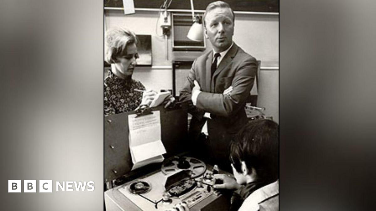 A sharply dressed man in a suit stands with his arms folded whilst a woman takes notes onto a a pad of paper. Another women sits in-front of an old fashioned tape reel player
