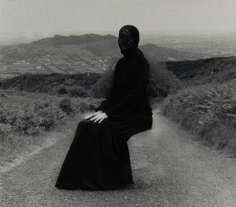 Órlaith Mac Eoin Manus   
Person squatting on forest path surrounded by greenery with hells visible in the background, wearing a dark floor-length outfit and lace-like head covering; face intentionally blurred for anonymity.
