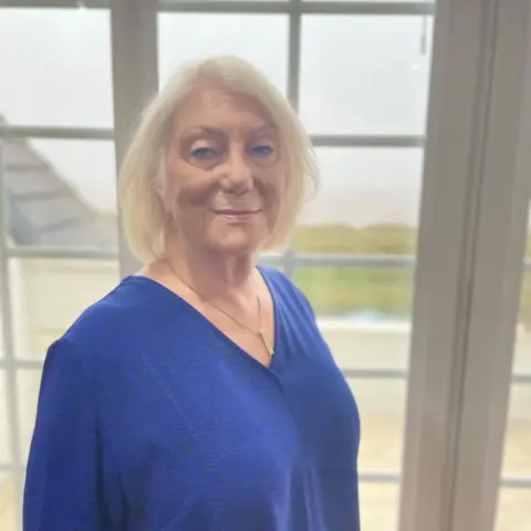 Carolyn is wearing a blue shirt and standing by her window at home looking to the camera. 