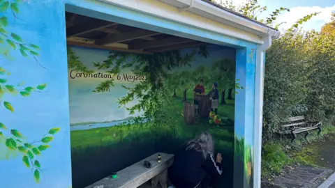 Claire Tagg We are looking at a bus shelter which has been transformed into a beautifully decorated scene of the countryside. In the corner a grey-haired woman is adding some touches of paint to the inside wall of the bus shelter