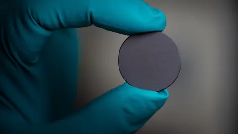 Tony Jolliffe/ BBC News A close-up of a gloved hand holding a small, circular grey disc - which is a sphere of material used in semiconductors. The glove is blue and the disc is being held between two fingers. 