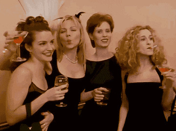 Four women in stylish dresses pose playfully with drinks at a social event, smiling and enjoying each other's company
