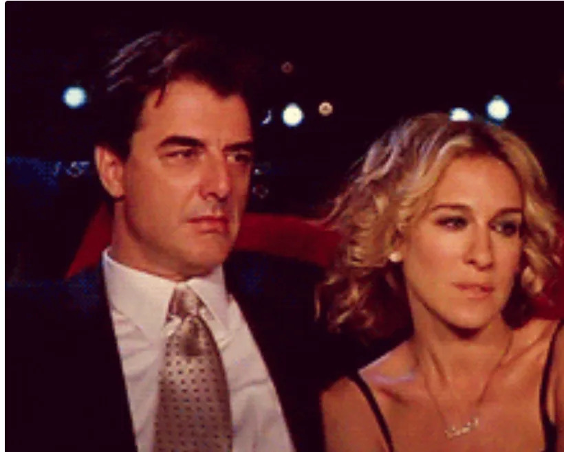 A man and woman sit closely together with serious expressions at an event, the woman wearing a strapless dress and her hair in loose curls