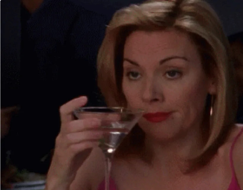 Person sitting, holding a martini glass, looking contemplative with a raised eyebrow, wearing a sleeveless top