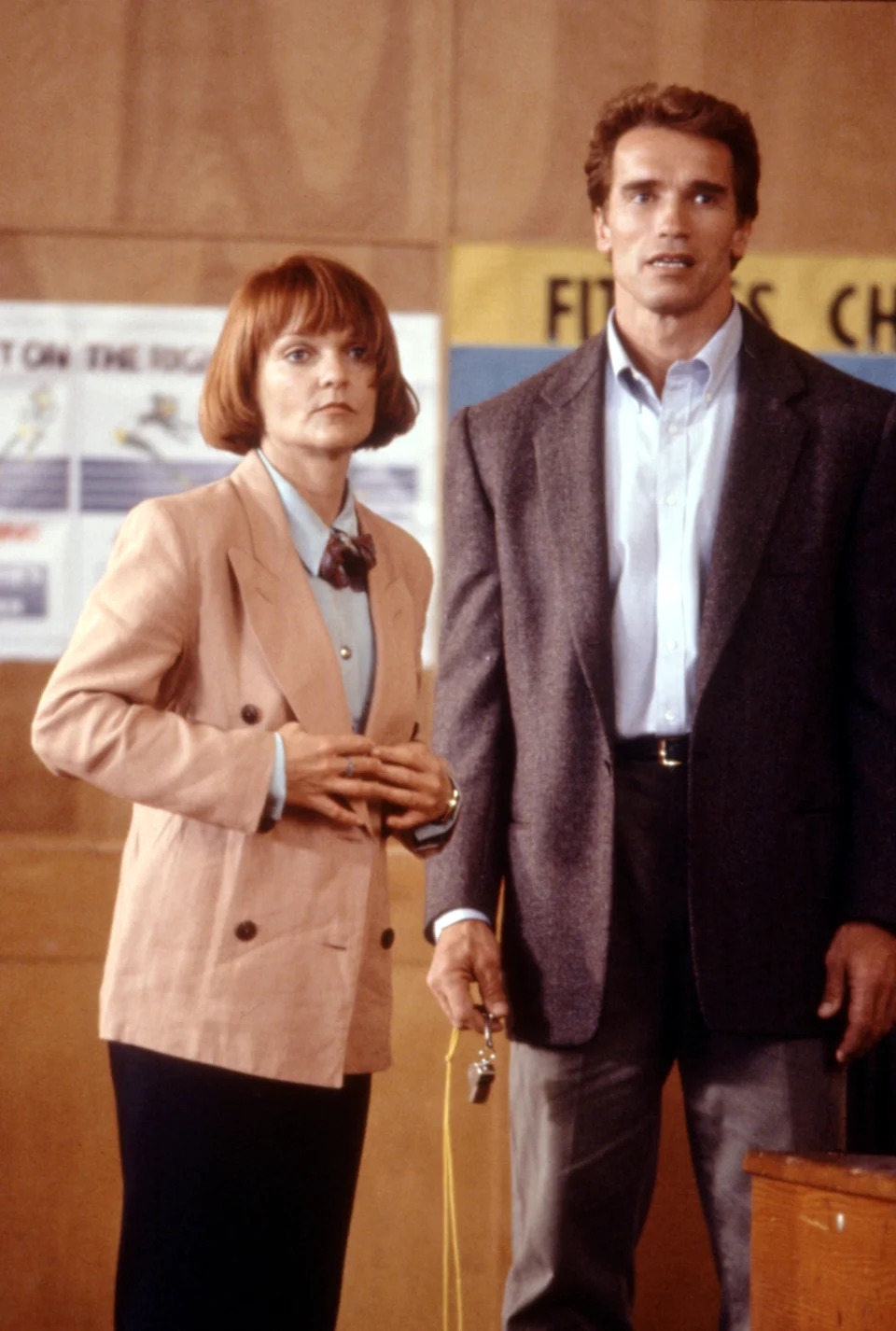 Two individuals stand indoors; one wears a blazer and tie, holding a whistle. The other wears a suit jacket, holding their hands together