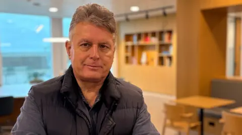 Ben Schofield/BBC A head and shoulders image of JP Allard in a modern-looking office area. He is wearing a dark gilet over a long sleeved casual blue shirt, which is open at the collar. He has relatively short, light coloured hair. Behind him, though out of focus in the picture, are chairs, tables and a book case.
