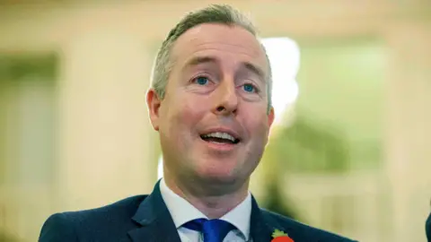 PA Media Paul Givan is wearing a navy blazer, white shirt, blue tie and a poppy. He has short grey hair. The background is blurred. 