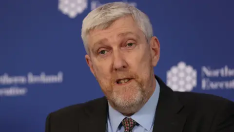 PA Media John O'Dowd is wearing a blue shirt, green tie and a black blazer. He has short grey hair and grey and red facial hair. Behind him is a blurred blue and white board displaying the Northern Ireland Executive logo. 