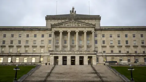 PA Media Stormont building from the front. The building is an off white colour. It has six columns on its front and steps leading to the doors. There is grass on either side of these steps. 