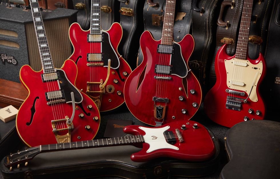 Five red electric guitars arranged with vintage cases and amplifiers in the background.