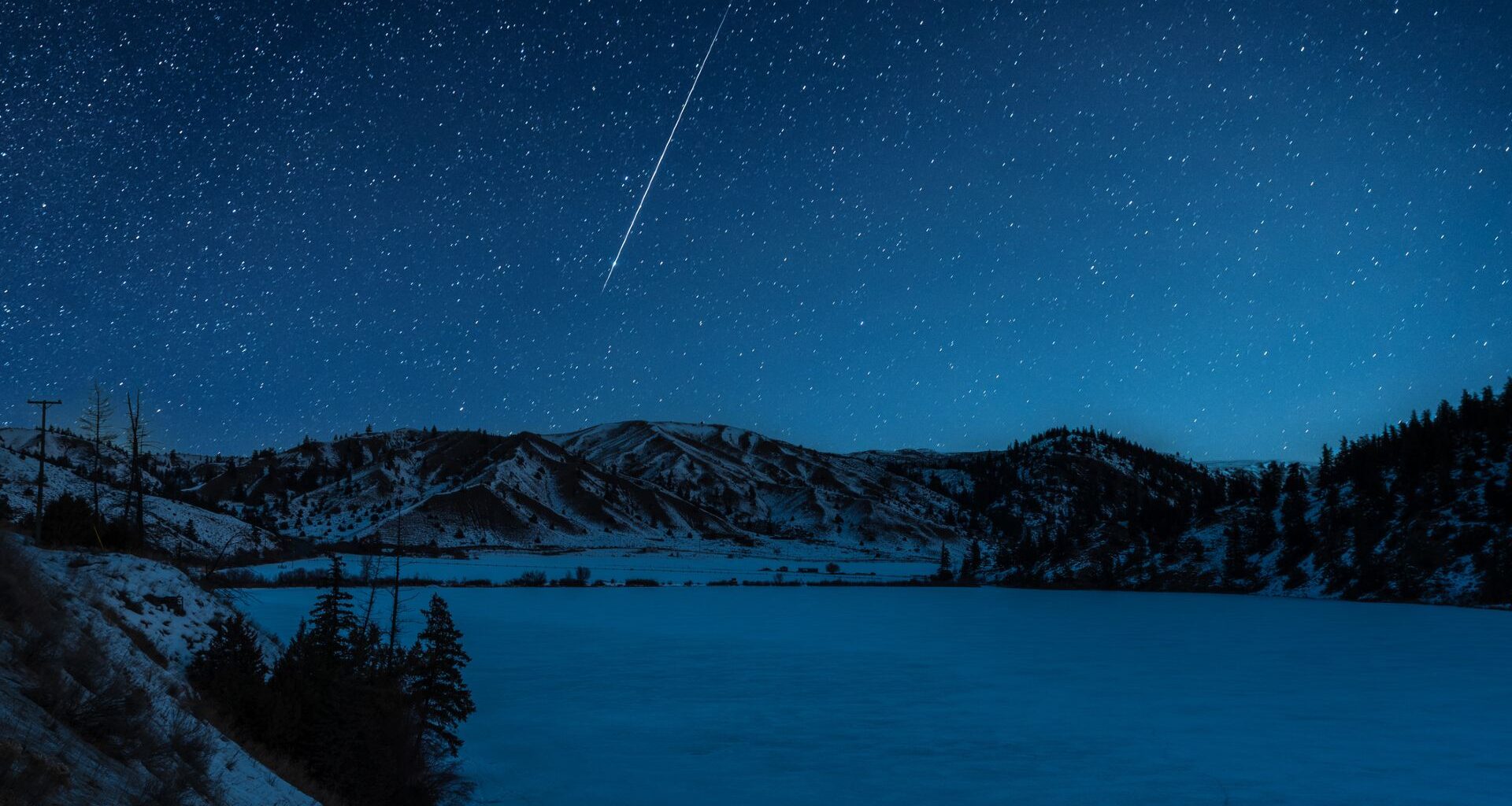 A shooting star is pictured streaking down through a starry sky above a frozen lake it night, bordered by snow-dappled hills.