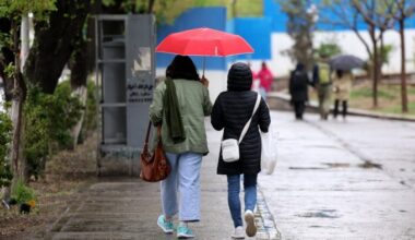 Two people walk side by side under a red umbrella on a wet sidewalk in Tehran in April as others walk in the background on a rainy day.