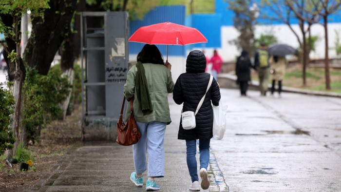 Two people walk side by side under a red umbrella on a wet sidewalk in Tehran in April as others walk in the background on a rainy day.