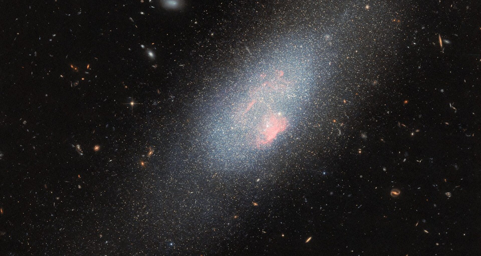 A pale blue dwarf galaxy on the black backdrop of space with some faraway galaxies. The galaxy itself resembles a fuzzy cloud of tightly packed stars, with a broad halo of stars dispersed around it. Spread across the galaxy’s core are several small, glowing patches of gas where there is a concentration of very hot stars.