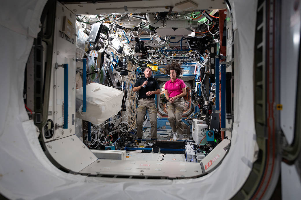 A man and a woman float inside the International Space Station. The man is wearing a dark shirt and holding a microphone. The woman wears a pink shirt, and her dark hair is standing straight up--there is no gravity to hold it down. Around them, the space station has white walls and many cables running along the walls and ceiling.