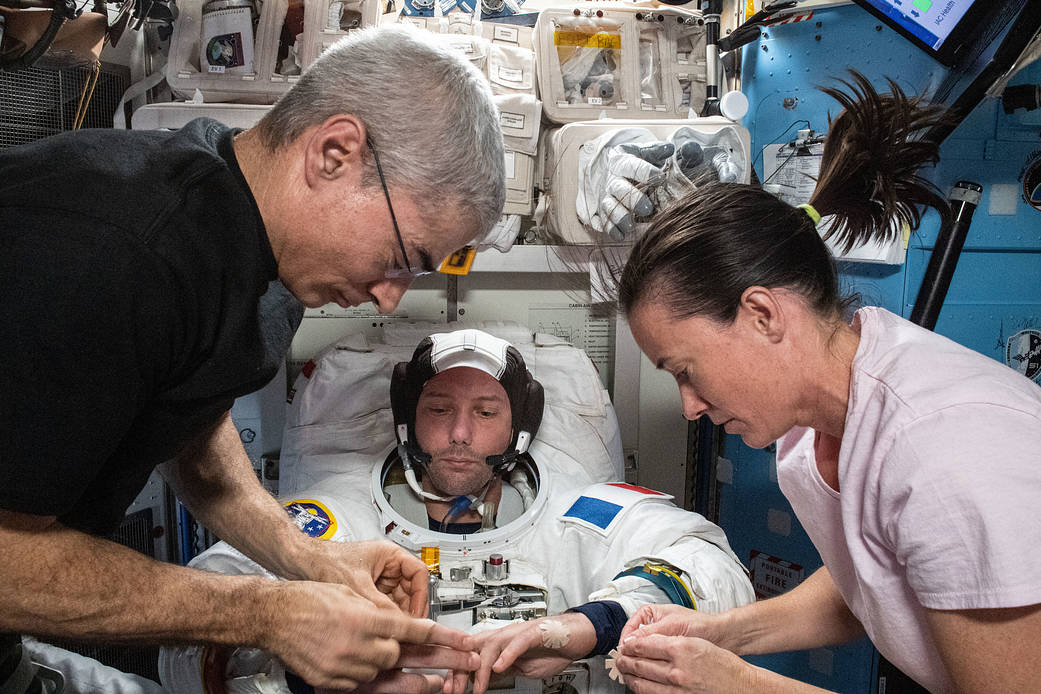 An astronaut is wearing a spacesuit in the center of the frame. He has a French flag patch on his left shoulder. The astronaut is not wearing a space helmet; he has a black and white cap on his head. On either side of him are two other astronauts, a man with gray hair and a woman with dark hair and a pink shirt. These two are inspecting the hands of the man in the spacesuit.