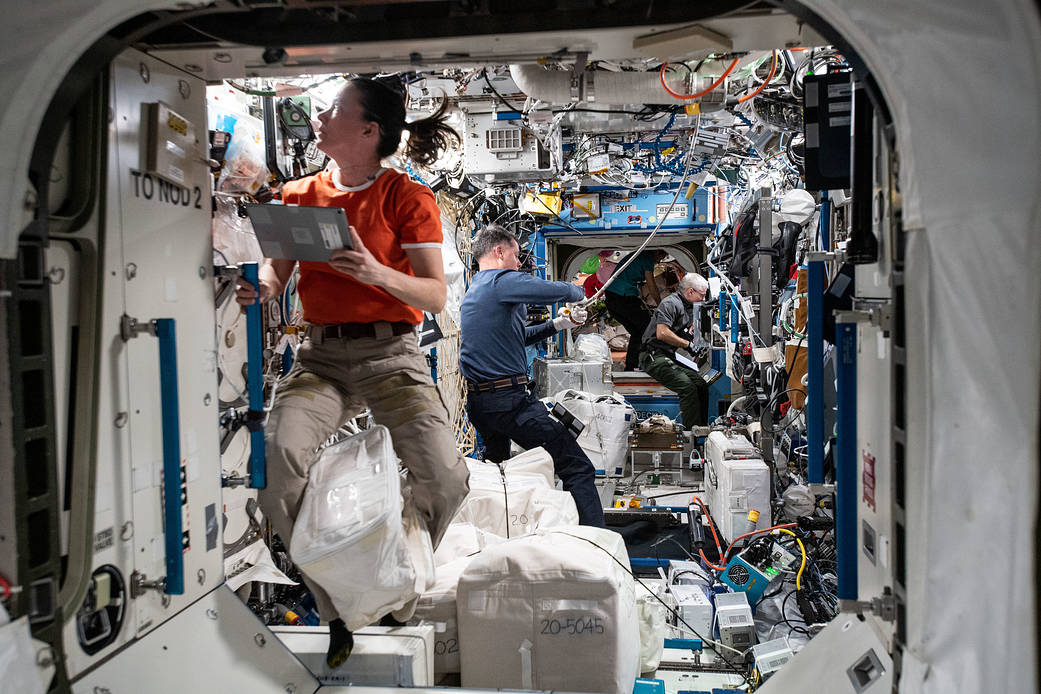 NASA astronauts work in the U.S. Destiny laboratory module. In the foreground, a woman with dark hair wearing an orange shirt looks intently at something. Behind her, a man in a blue shirt with dark hair adjusts something connected to the ceiling. In the background, another man appears busy.