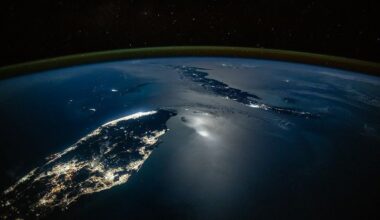 An oblique nighttime photo shows the Florida Peninsula on the left and Cuba on the middle-right, with clusters of city lights illuminating both landmasses. Moonglint shimmers across part of the ocean surface, while green airglow and the darkness of space stretch across the top of the image.