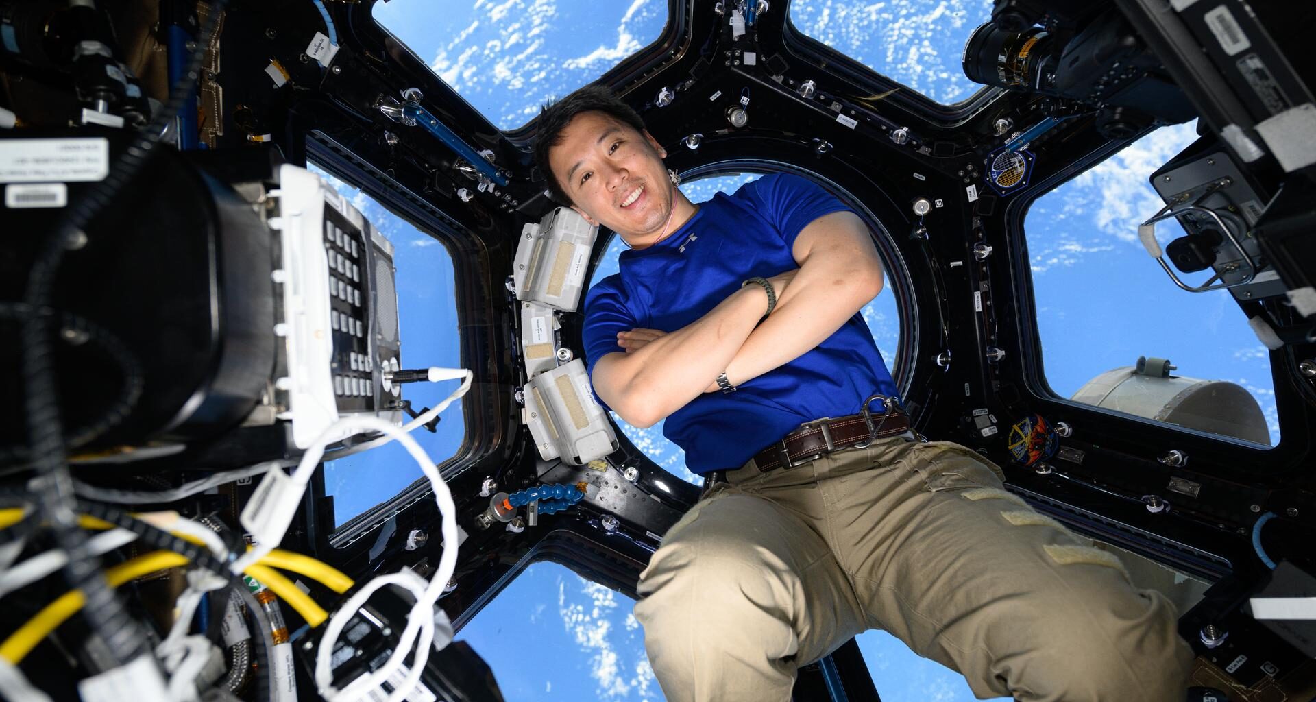 NASA astronaut Jonny Kim floats in the center of the image with his arms crossed, smiling. He wears a blue shirt and khaki pants. Behind him, the seven windows of the space station’s cupola reveal Earth’s blue oceans below. In the foreground, on the left, a rectangular device with a keypad and multiple wires is visible.