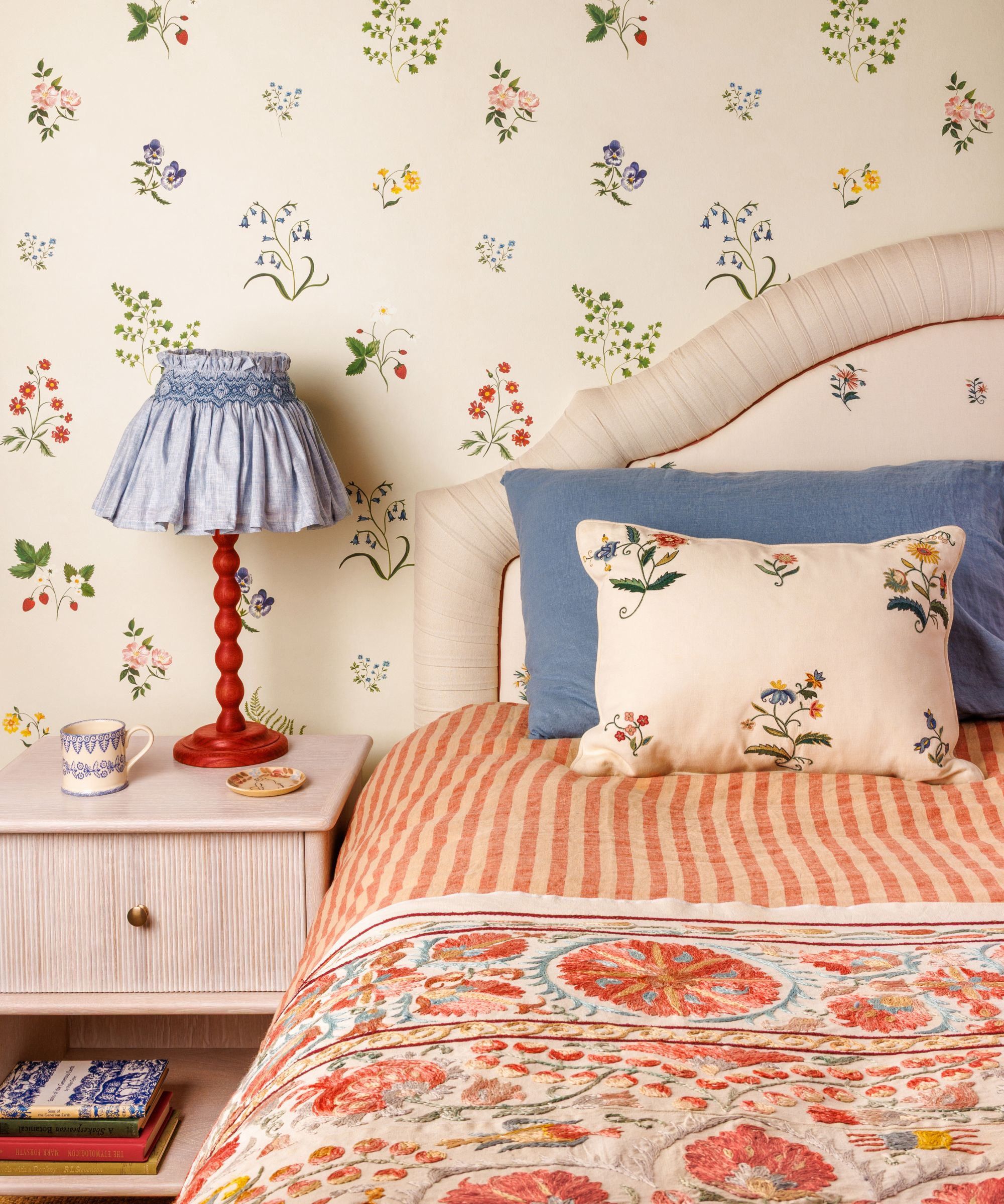 A charming, patterned bedroom featuring delicate floral wallpaper and a bed with a fan-shaped cream headboard. The bedding includes an orange striped duvet and an ornate floral embroidered quilt, paired with a red-based lamp with a pleated blue fabric shade on a light wood nightstand