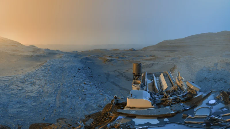 A panoramic view from the surface of Mars shows rocky, barren terrain under a gradient sky, transitioning from orange to blue, with part of a rover and its equipment visible in the foreground.