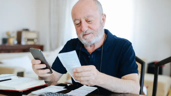 Senior man looking at bills and using his smartphone to budget and plan finances.