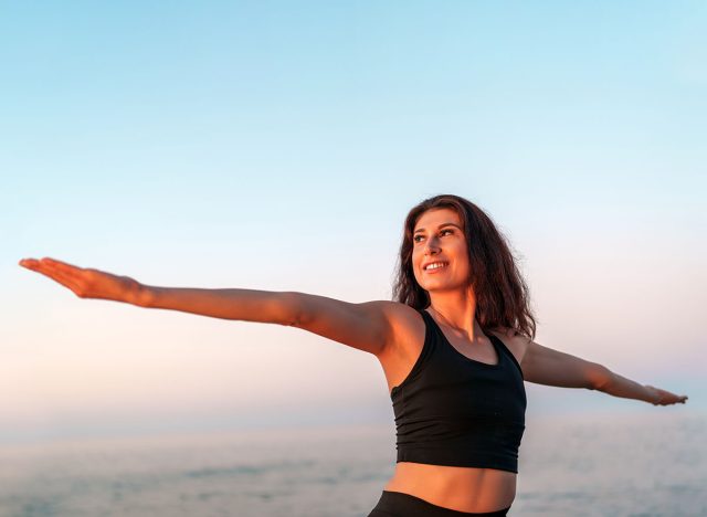 Smiling sportswoman practicing yoga on the beach at sunset, arms outstretched and embracing the serene beauty of the ocean