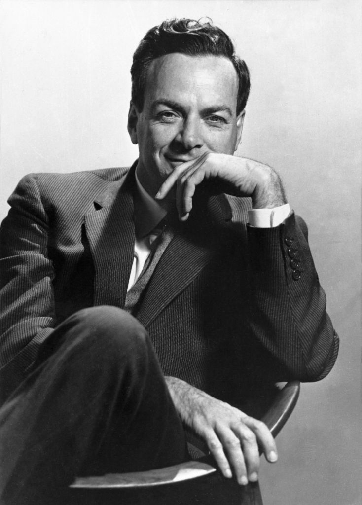 Black and white professional headshot of Richard Feynman. He sits in a chair facing the camera, with his knee propped up on the chair and his hand partially covering his mouth.