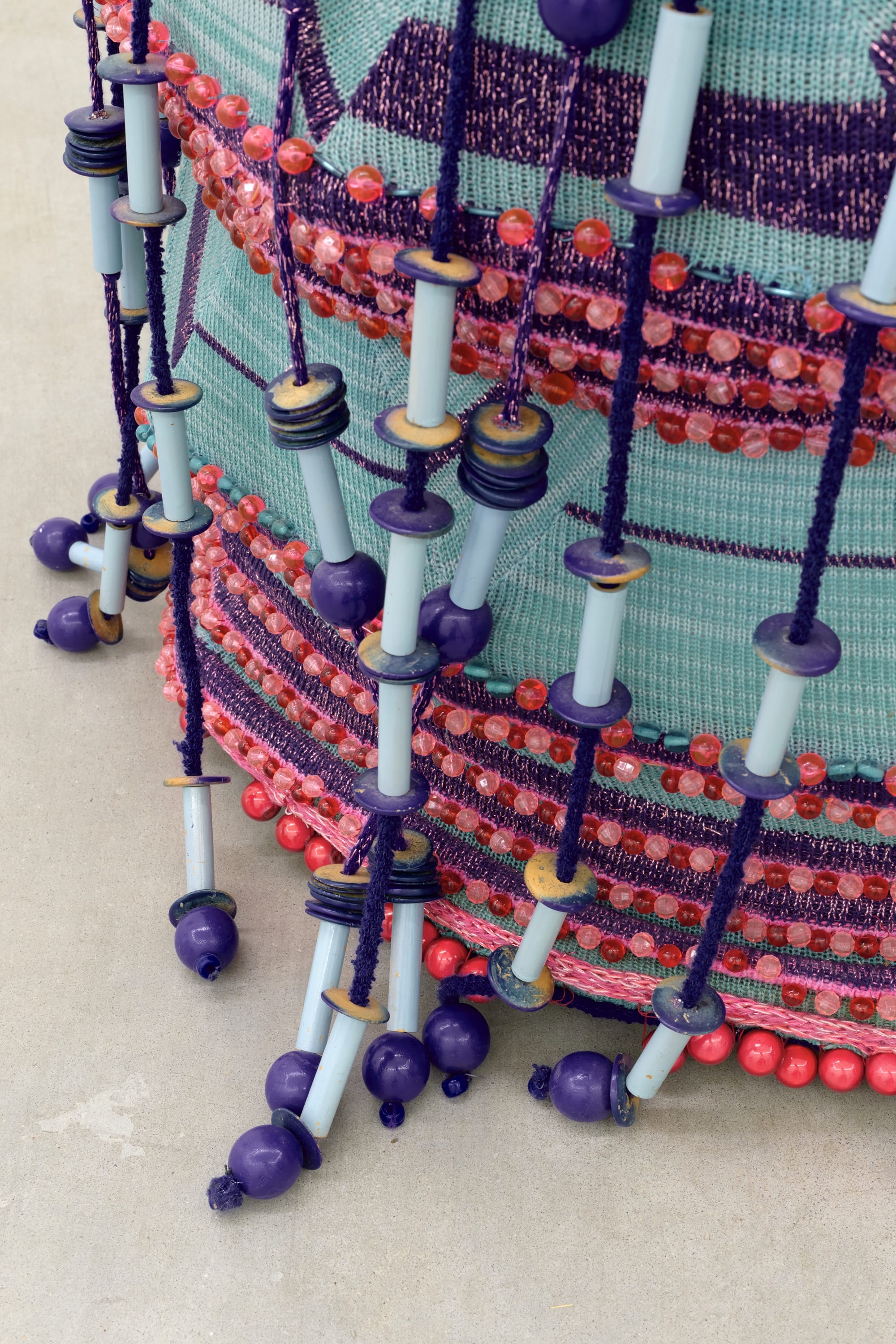 A detail of a woven textile sculpture by Jim Drain loosely resembling a large teal and magenta basket with a lid and many long strands or tassels