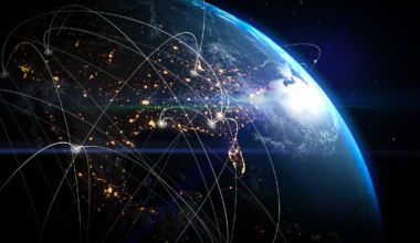View of Earth from space at night showing illuminated city lights and glowing data network lines connecting various points across continents, symbolizing global communication, technology, and interconnected digital infrastructure. The image highlights the curvature of the planet with a bright sunrise over the horizon and deep space background.