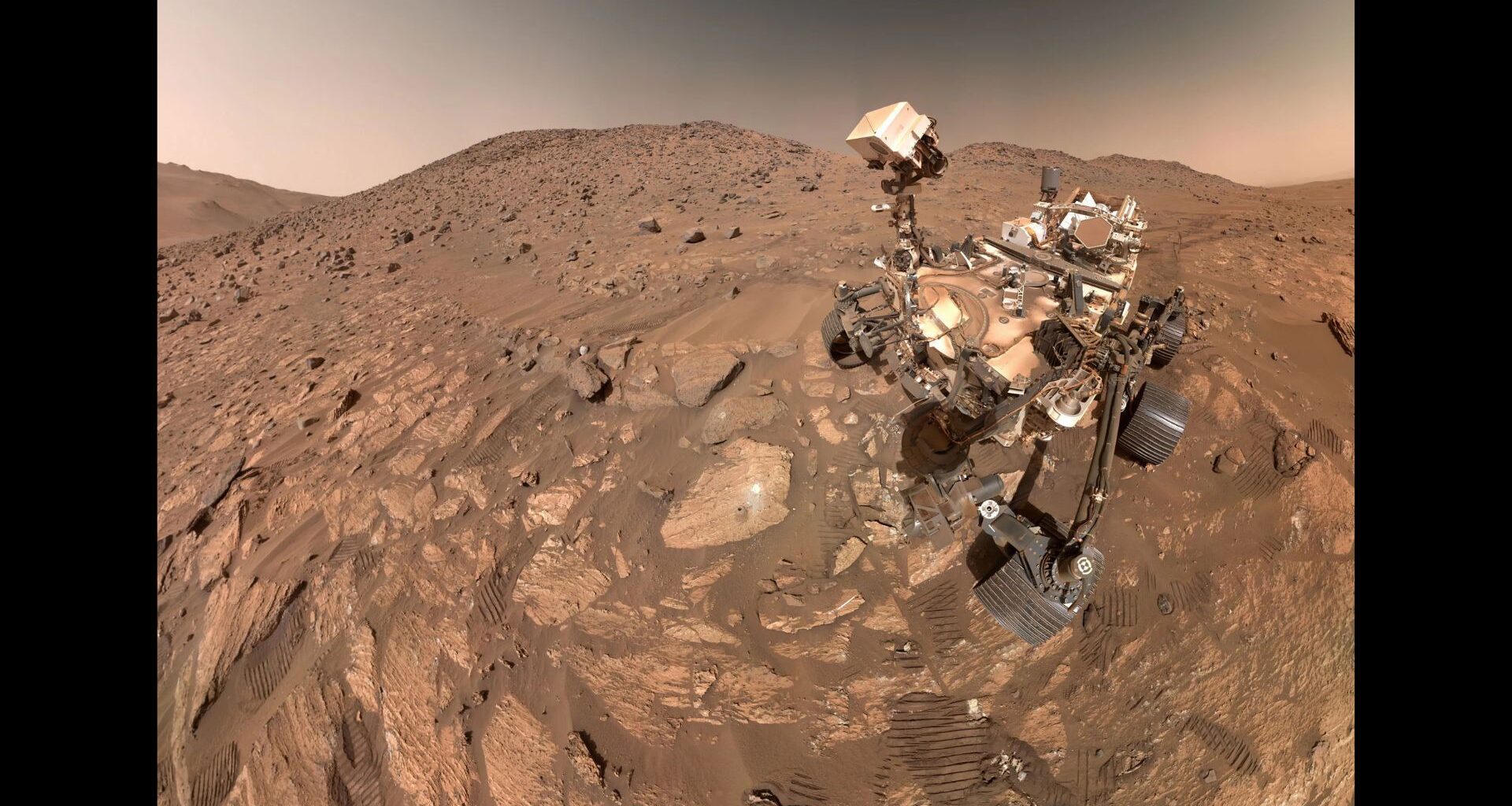 Photo of the Perseverance rover taken by the rover from above. The Martian landscape makes up most of the photo&mdash;reddish brown rocks and dirt.