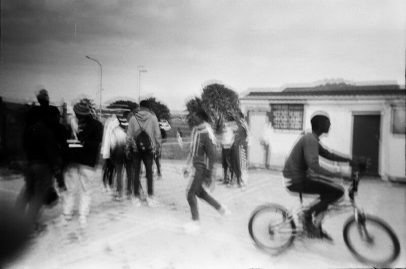 A black-and-white photo shows a group of people walking together and one person riding a bicycle in the foreground. The image is blurry, giving a sense of motion. Buildings and streetlights are visible in the background.