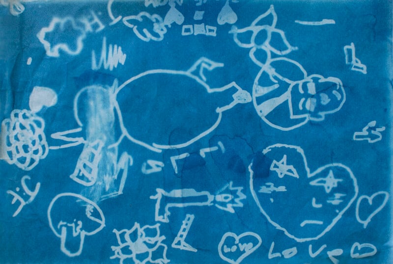 Blue paper with white, abstract and childlike drawings, including a heart labeled "Love," a sad face, a flower, a gun, a mushroom, stars, hearts, and various shapes and scribbles scattered throughout.