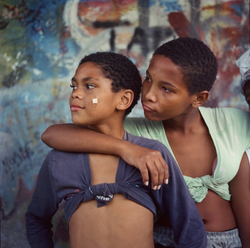 Two children stand together against a colorful, graffiti-covered wall. One has an arm around the other’s shoulders, who has a bandage on their cheek. Both are wearing shirts tied in the front and look off to the side.