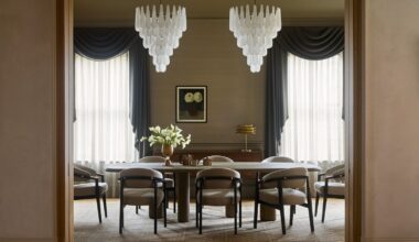 A symmetrical, sophisticated dining room viewed through a doorway. The space features a long wooden table surrounded by curved grey armchairs, two large tiered glass chandeliers, and tall windows with dramatic charcoal grey swags and sheer curtains