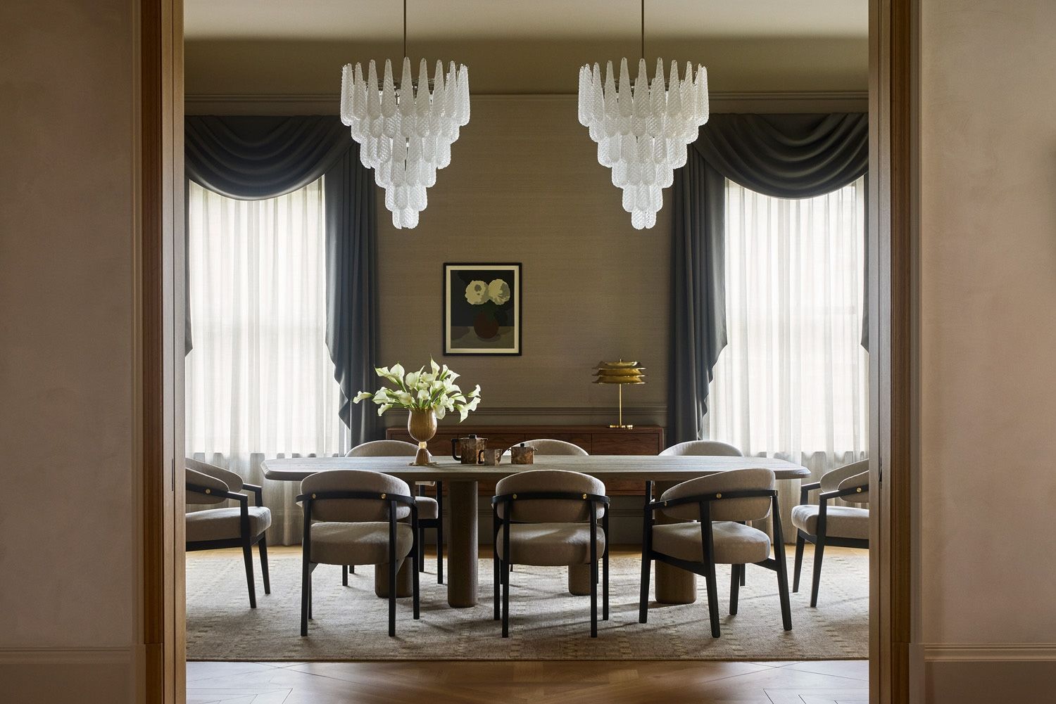 A symmetrical, sophisticated dining room viewed through a doorway. The space features a long wooden table surrounded by curved grey armchairs, two large tiered glass chandeliers, and tall windows with dramatic charcoal grey swags and sheer curtains
