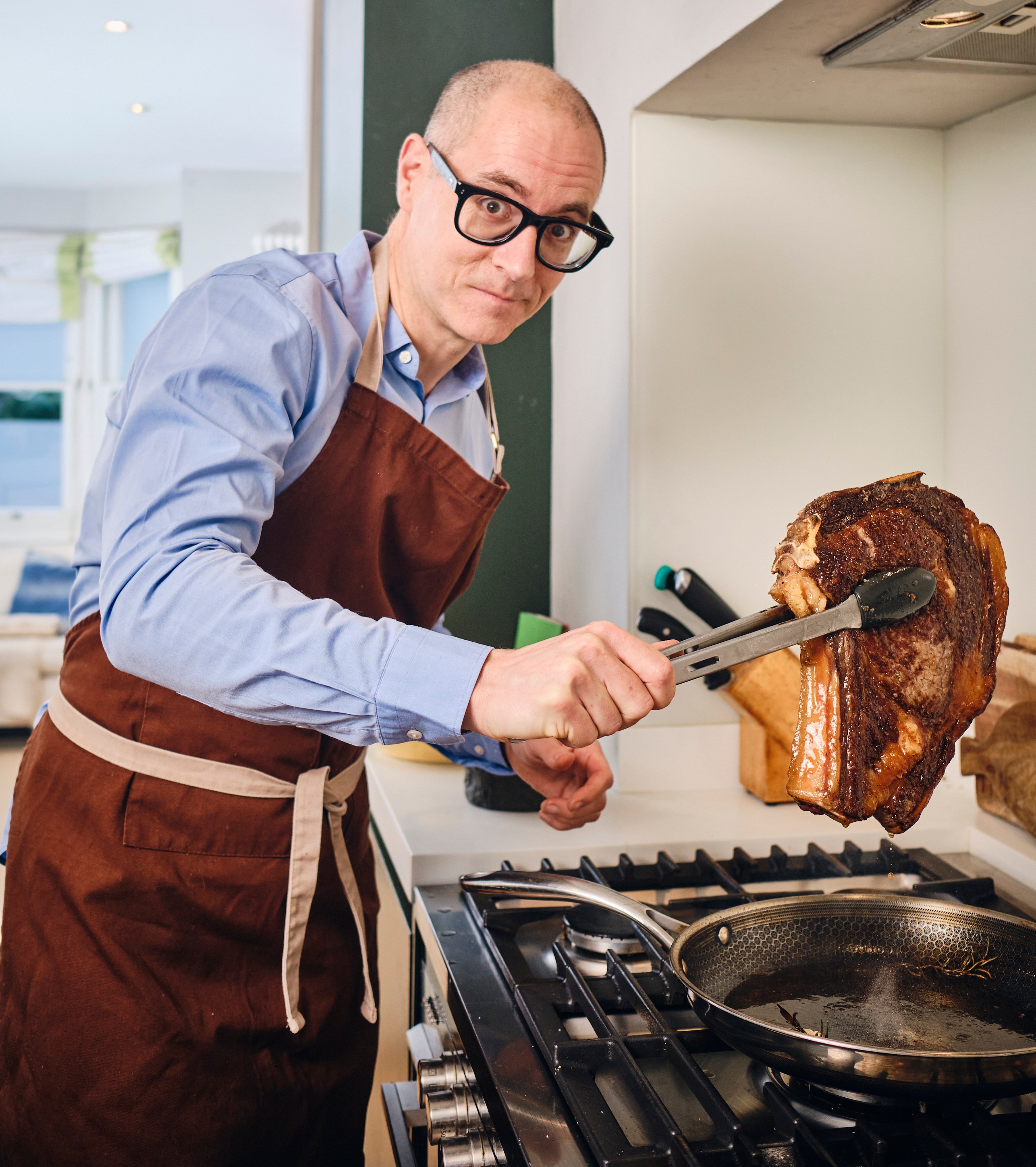 Harry Wallop holding a large cut of meat with tongs over a frying pan.